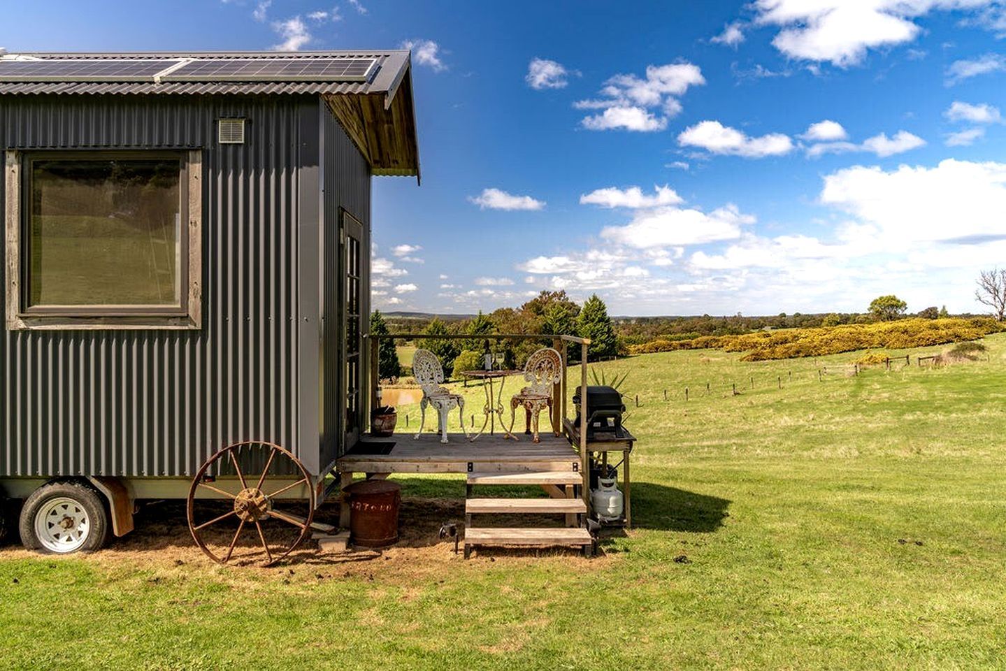A small, single-story metal building with a porch, a vintage wheel, and a table with chairs.