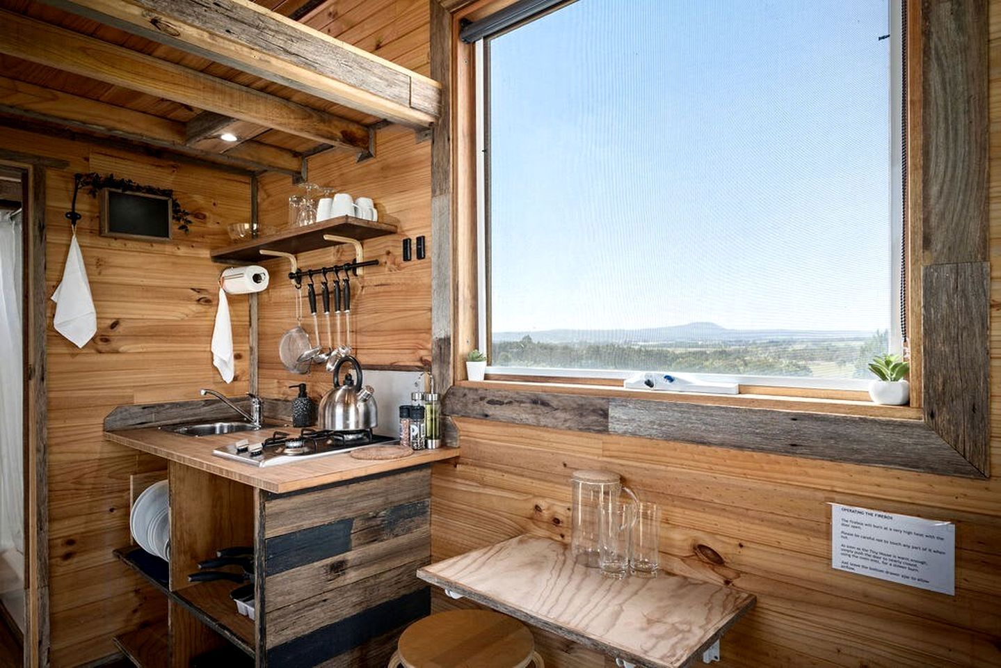 A rustic kitchen with wooden walls, a window with a view, and basic amenities like a kettle and utensils.