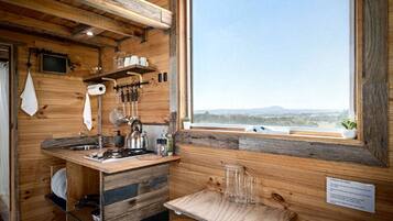 A rustic kitchen with wooden walls, a window with a view, and basic amenities like a kettle and utensils.