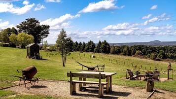 A picnic area with a wooden bench, table, and chairs, surrounded by a grassy field and a small shed.