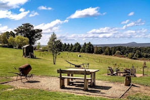 A picnic area with a wooden bench, table, and chairs, surrounded by a grassy field and a small shed.