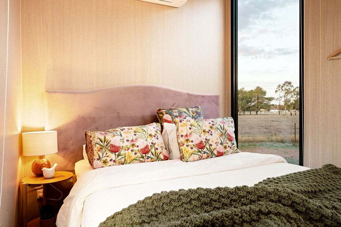 Un dormitorio con cama, almohadas, una lámpara y vistas al exterior.