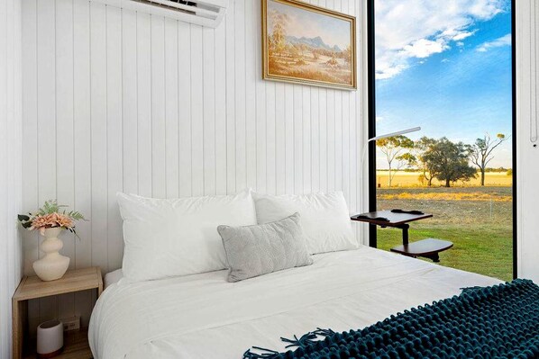 A bedroom with a large bed, a bedside table with a vase of flowers, and a view of a grassy field through a window.
