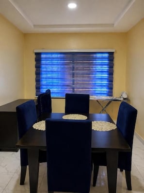 Un comedor con una mesa, sillas, una ventana con persianas y piso de baldosas.