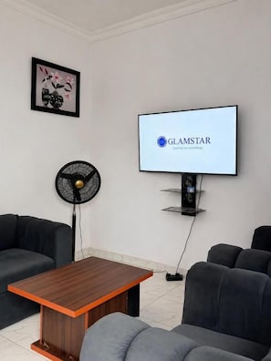 Una sala de estar moderna con un televisor de pantalla plana mostrando 'GLAMSTAR' en la pantalla, un ventilador negro y una mesa de centro de madera.