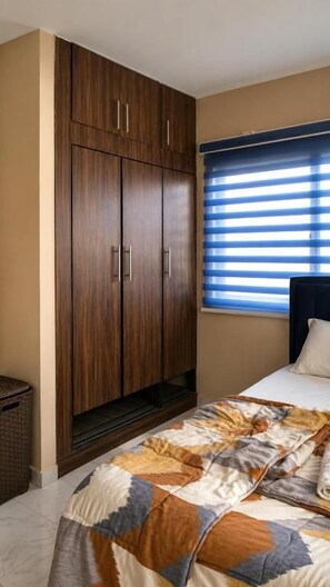 Een slaapkamer met een bed, een houten garderobe, een raam met jaloezieën en een bed met een decoratieve spreideken.