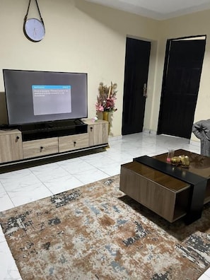 Uma sala de estar com televisão de tela plana sobre suporte de madeira, mesa de centro, tapete estampado e um vaso com flores.