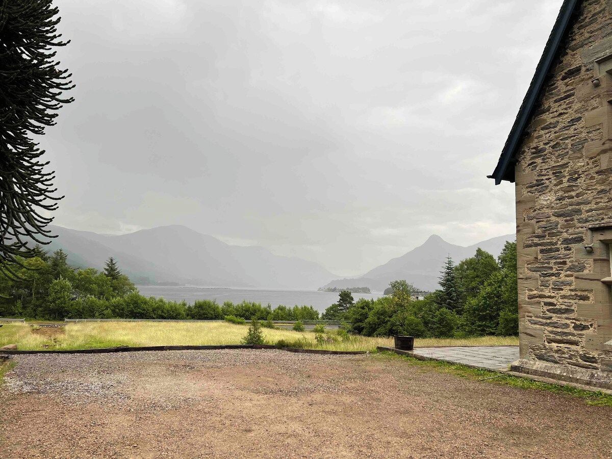 A gravel driveway leading to a stone building with a view of mountains and a body of water in the distance.