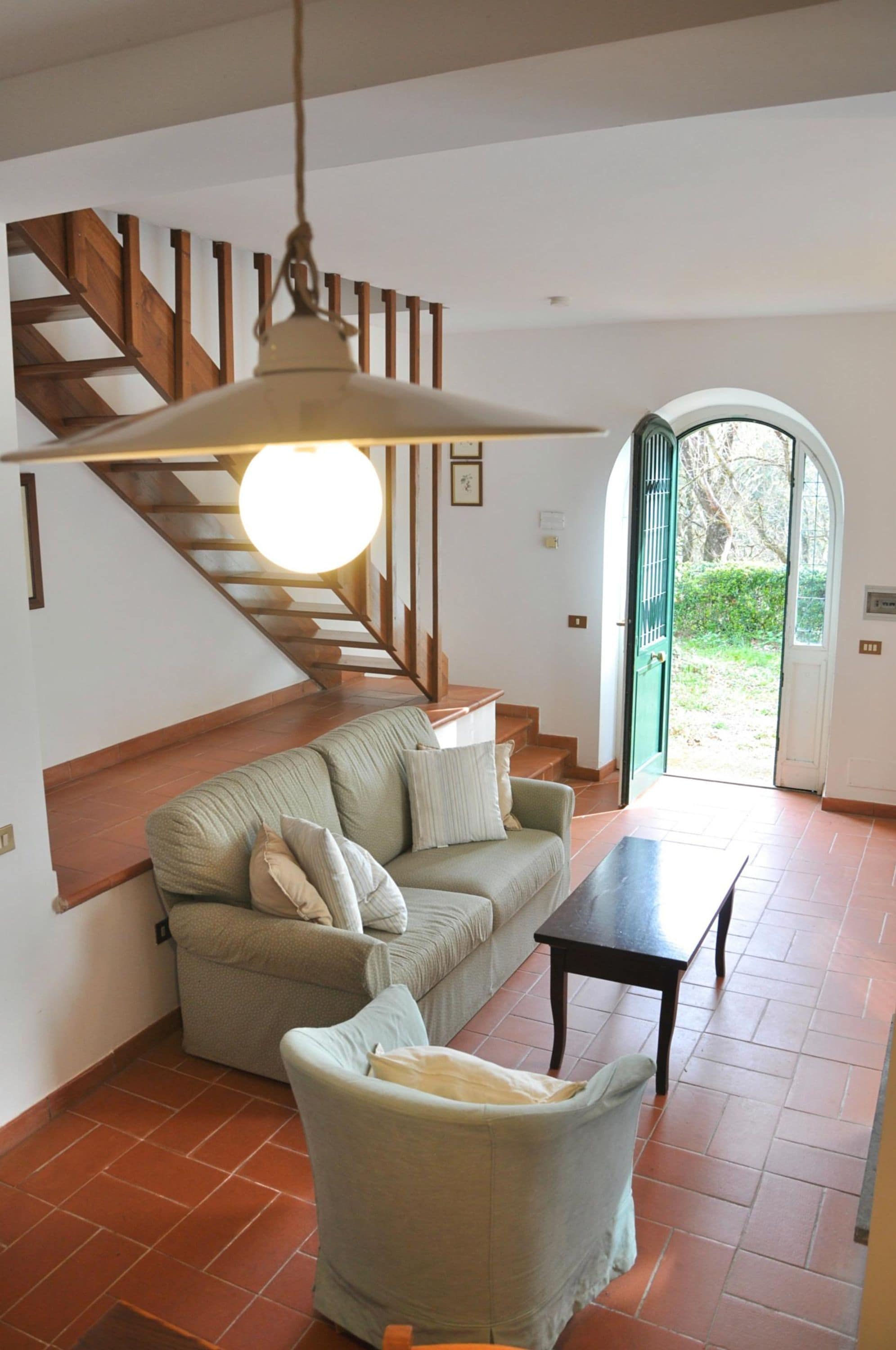 A living room with a sofa, armchair, and a table. There is a staircase with wooden railings and a hanging light fixture.