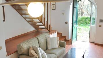 A living room with a sofa, armchair, and a table. There is a staircase with wooden railings and a hanging light fixture.