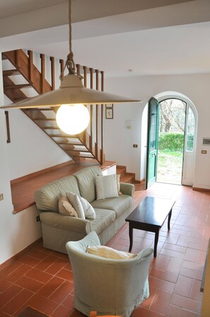 A living room with a sofa, armchair, and a table. There is a staircase with wooden railings and a hanging light fixture. - Montecasciano Margherita (Capranica)