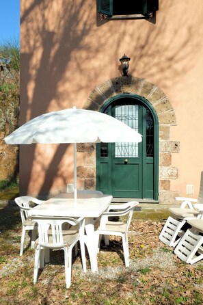 A patio with a table and chairs, an umbrella, and a building with a green door and stone architecture. - Montecasciano Margherita (Capranica)