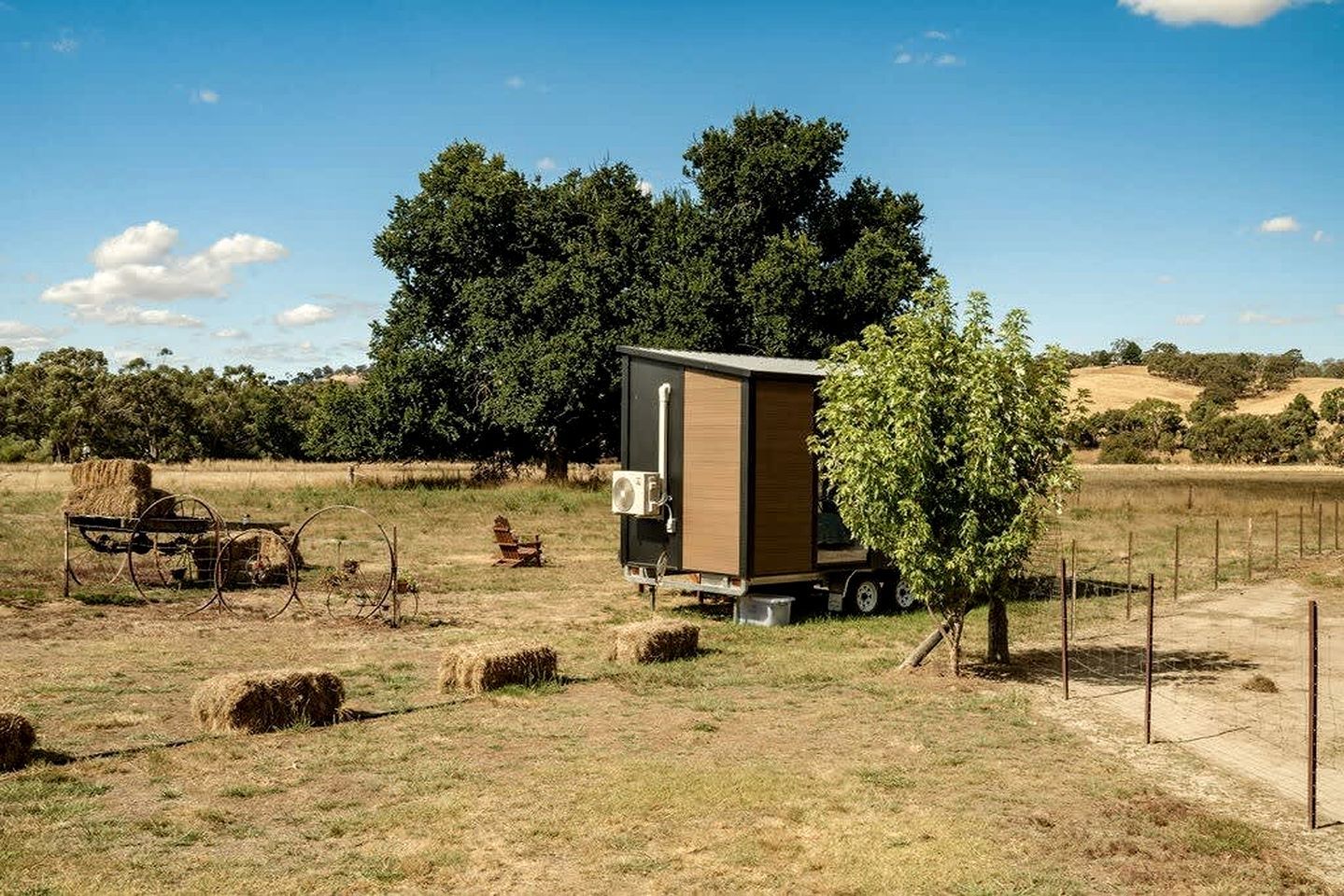 A mobile home with a large window, situated in a rural setting with hay bales and a tree nearby.