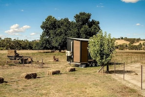 A mobile home with a large window, situated in a rural setting with hay bales and a tree nearby.