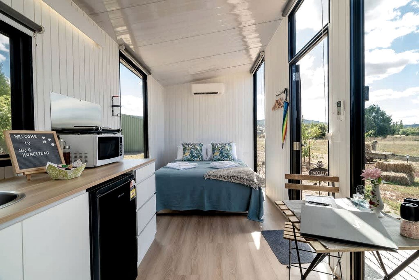 A compact, modern room with a bed, a small kitchenette, and a large window offering a view of the countryside.