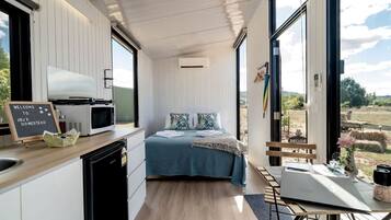 A compact, modern room with a bed, a small kitchenette, and a large window offering a view of the countryside.