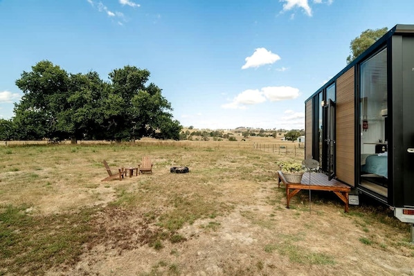 Une maison mobile moderne dotée d’une grande fenêtre, d’une terrasse et d’un terrain herbeux avec des arbres en arrière-plan.
