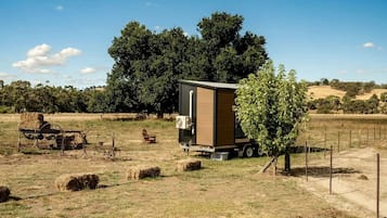 A mobile home with a large window, situated in a rural setting with hay bales and a tree nearby.