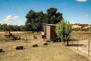 A mobile home with a large window, situated in a rural setting with hay bales and a tree nearby.