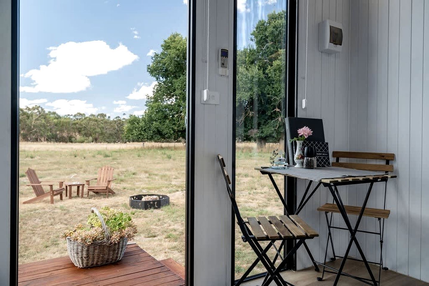 A room with a view of a grassy field, a wooden table with chairs, and a small garden outside.