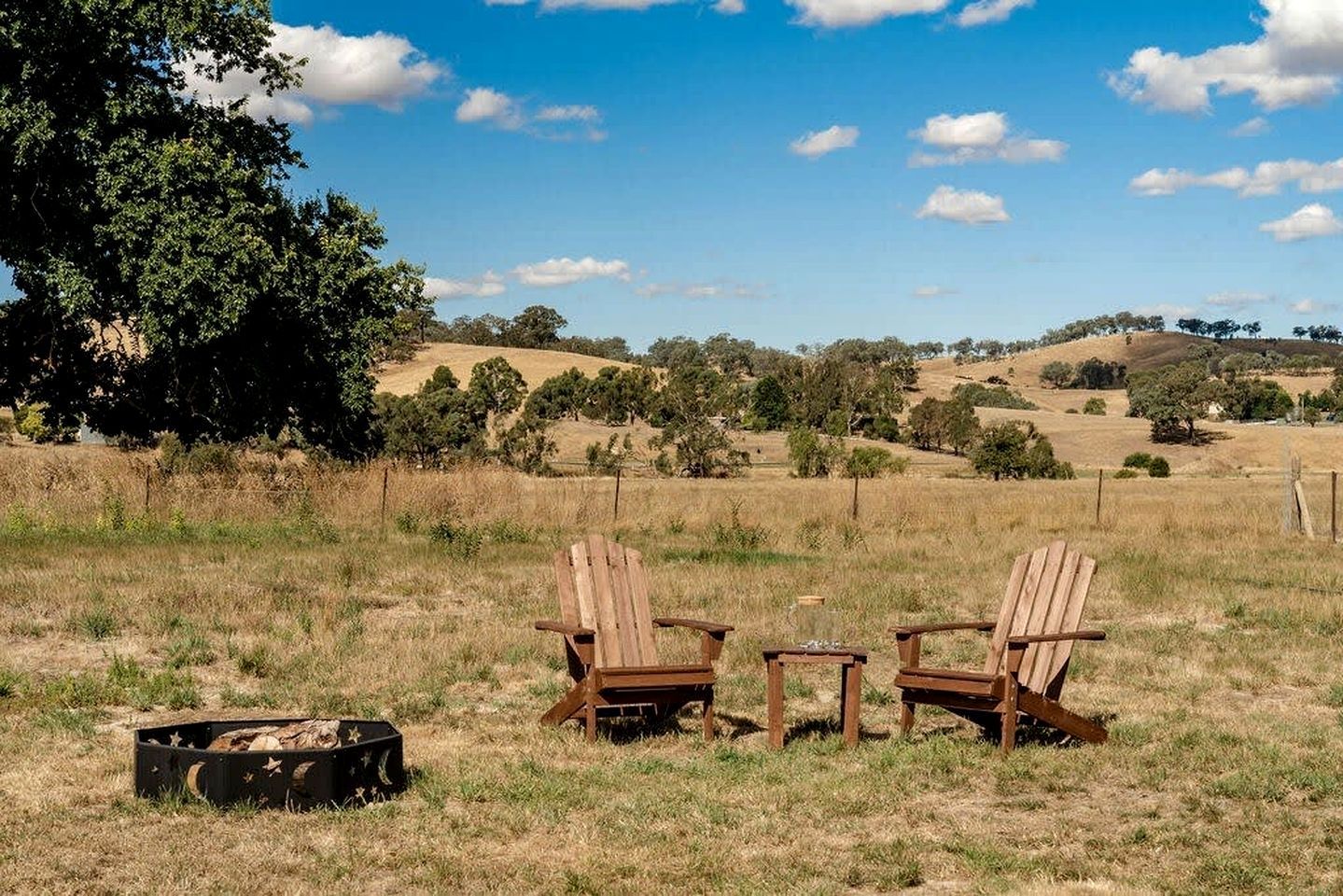 Two wooden chairs in a grassy field with a tree and hills in the background.