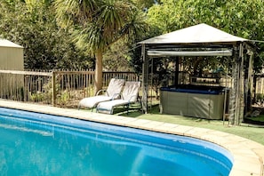 A swimming pool with lounge chairs and a hot tub, surrounded by a wooden fence and trees.