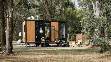 A modern, compact cabin with a black and wooden exterior, situated in a wooded area with a fire pit and outdoor seating.