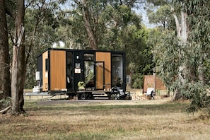 A modern, compact cabin with a black and wooden exterior, situated in a wooded area with a fire pit and outdoor seating.