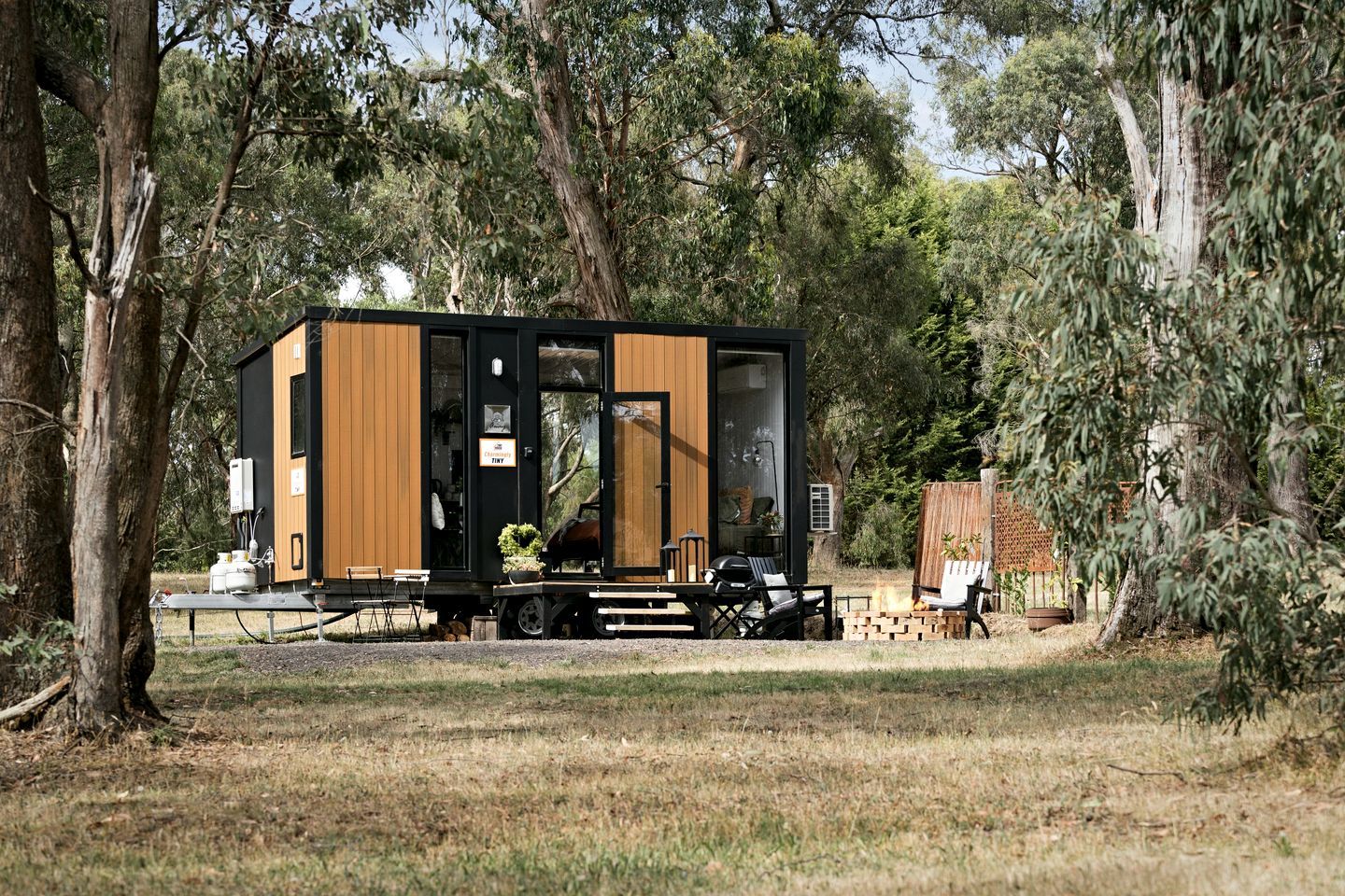 A modern, compact cabin with a black and wooden exterior, situated in a wooded area with a fire pit and outdoor seating.