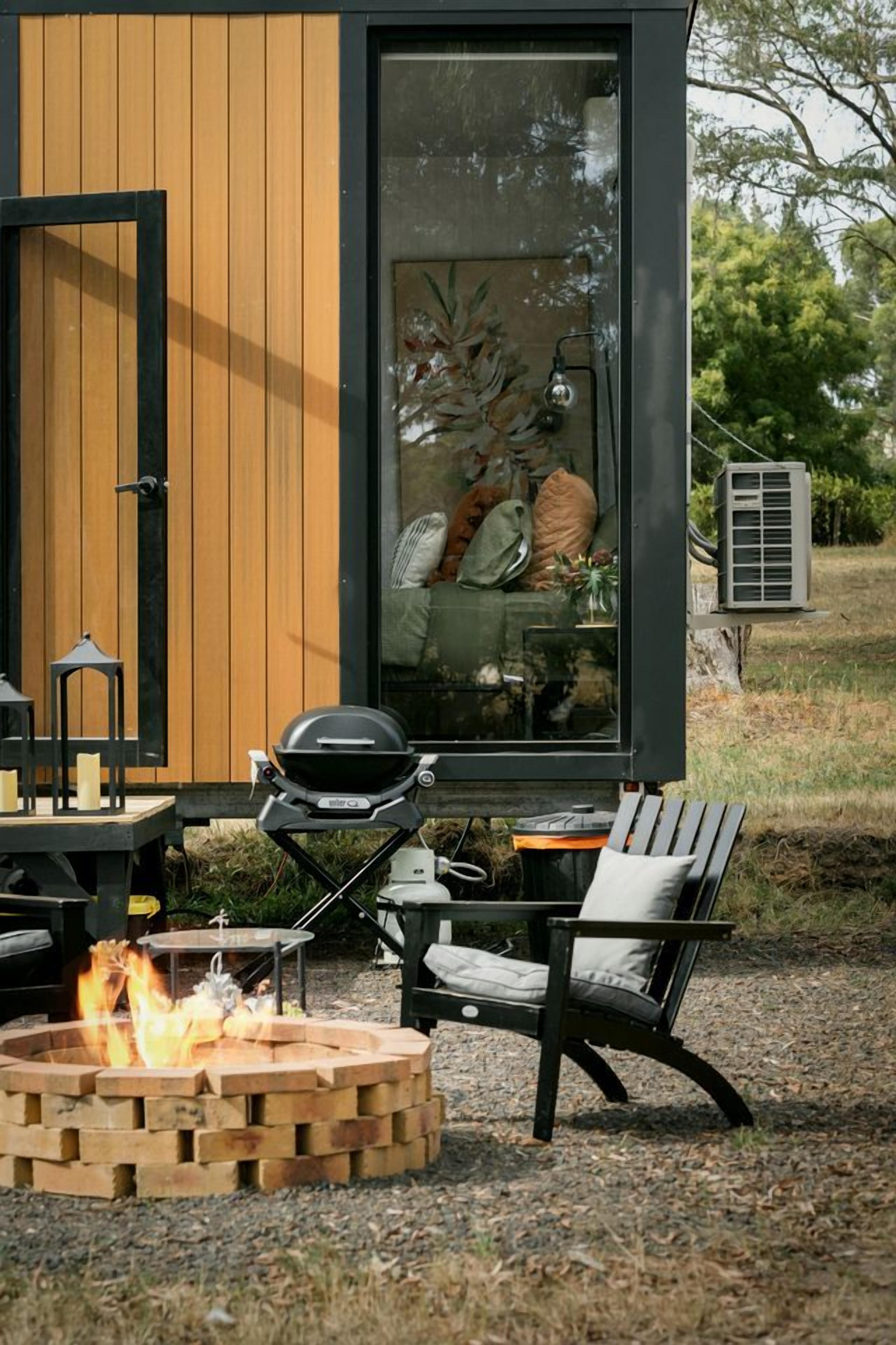 A modern outdoor seating area with a fire pit, a grill, and a view of a cozy interior.