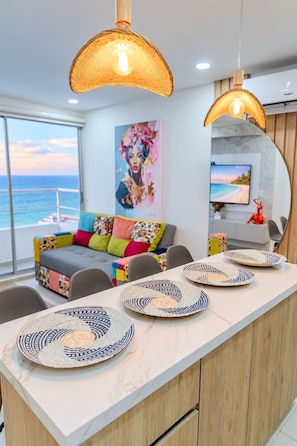 A modern living room with a sofa, colorful cushions, a dining area with a long table and plates, and a large painting on the wall. - Ocean Bloom | Sea Views, Beach, 3BR & Pool (Cartagena)