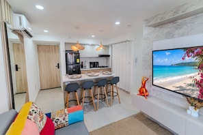 A modern living room with a sofa, a TV displaying a beach scene, and a kitchen area with a refrigerator, stove, and bar stools. - Ocean Bloom | Sea Views, Beach, 3BR & Pool (Cartagena)