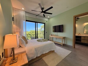 A bedroom with a bed, bedside table, lamp, ceiling fan, TV, and a view of trees through the window. - Casa Pura Aventura | Pool, Views & Coastal Luxury (Playa Hermosa)