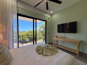 A bedroom with a large bed, a TV mounted on the wall, and a balcony with a view of greenery. - Casa Pura Aventura | Pool, Views & Coastal Luxury (Playa Hermosa)
