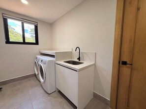 A compact laundry room with a washing machine, a sink, and a window with a view of greenery. - Casa Pura Aventura | Pool, Views & Coastal Luxury (Playa Hermosa)