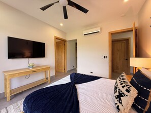 A bedroom with a bed, a TV mounted on the wall, a ceiling fan, an air conditioning unit, and wooden doors. - Casa Pura Aventura | Pool, Views & Coastal Luxury (Playa Hermosa)