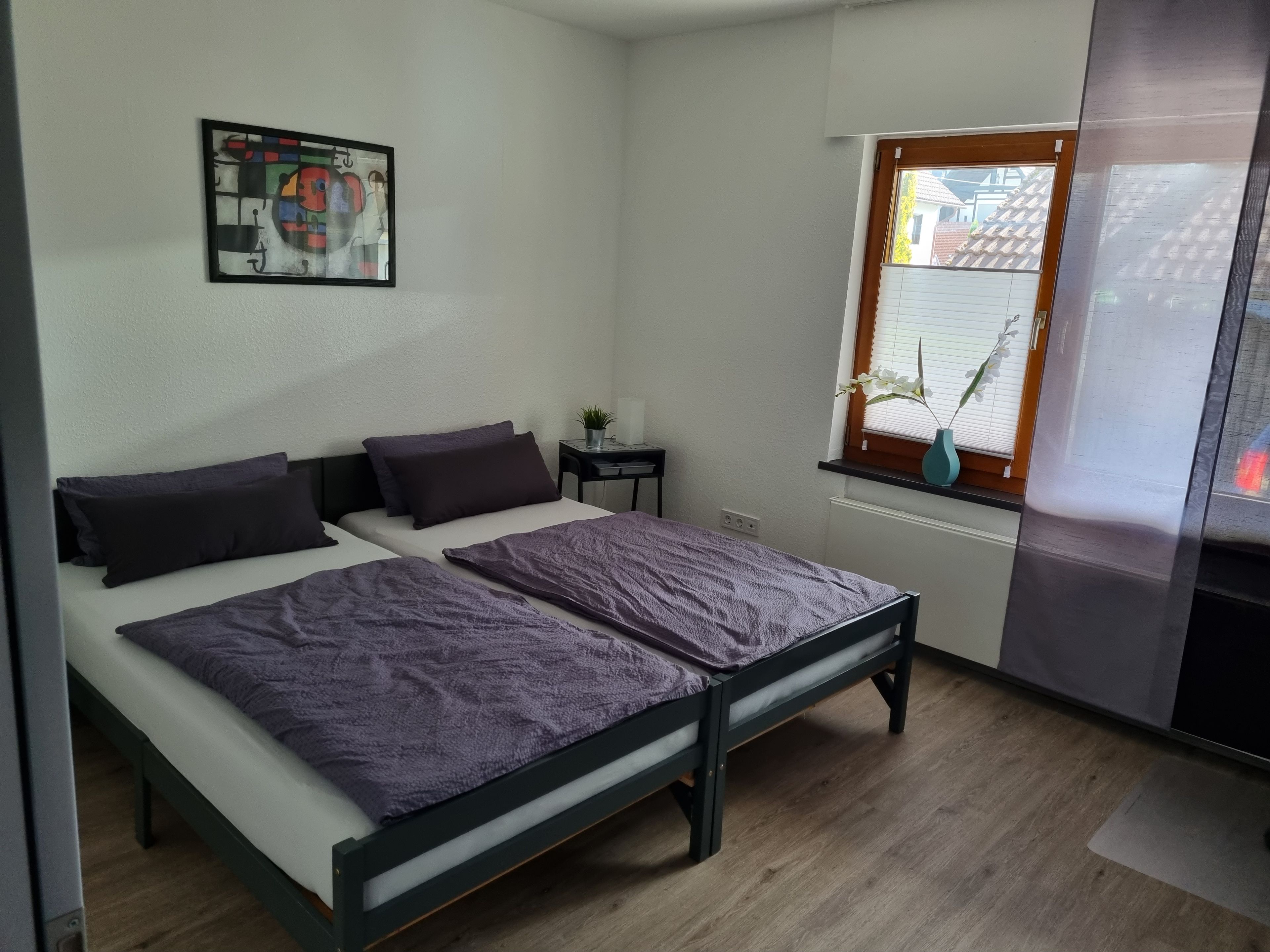 Ein Zimmer mit einem Bett, einem kleinen Tisch, einem Fenster mit Jalousien und einem Wandbild.