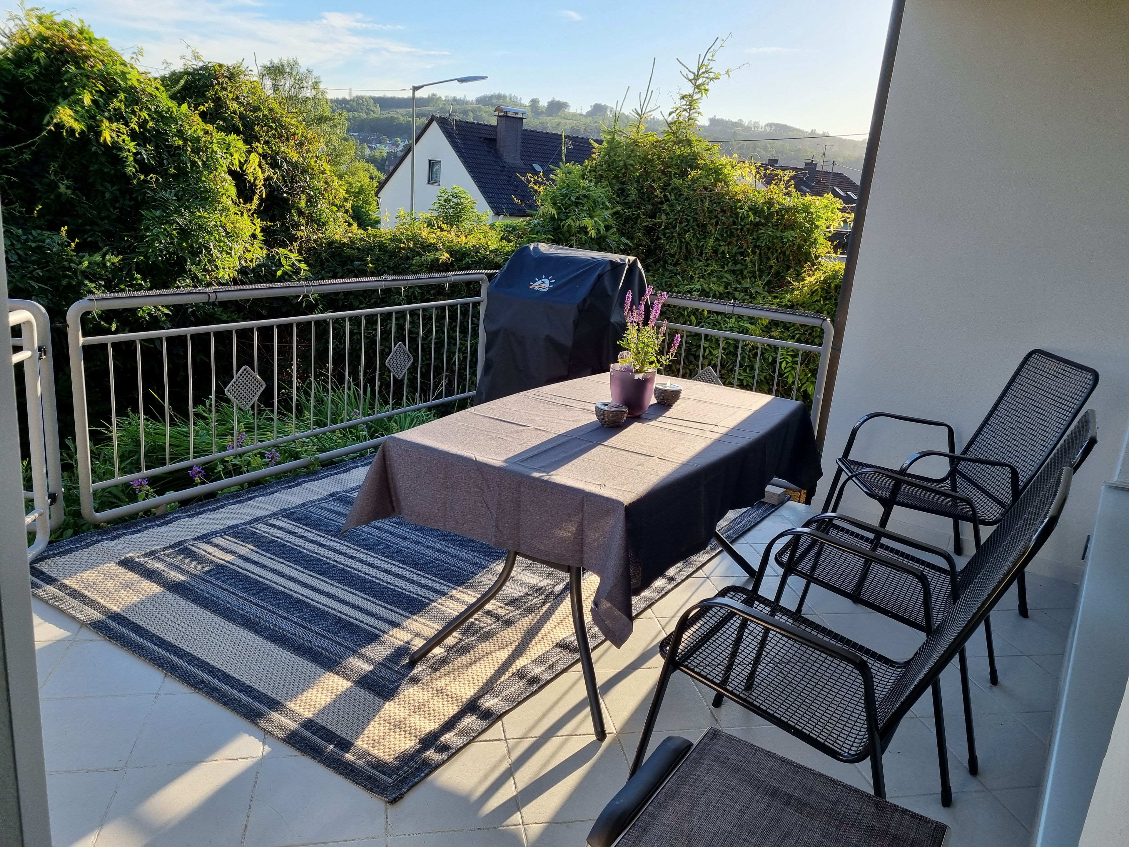 Ein Balkon mit Tisch und Stühlen, einem Blumentopf und Blick auf die umliegende Bepflanzung und Gebäude.