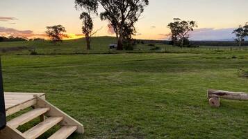 A grassy field with a wooden staircase leading up to a building, surrounded by trees and a sunset in the background.