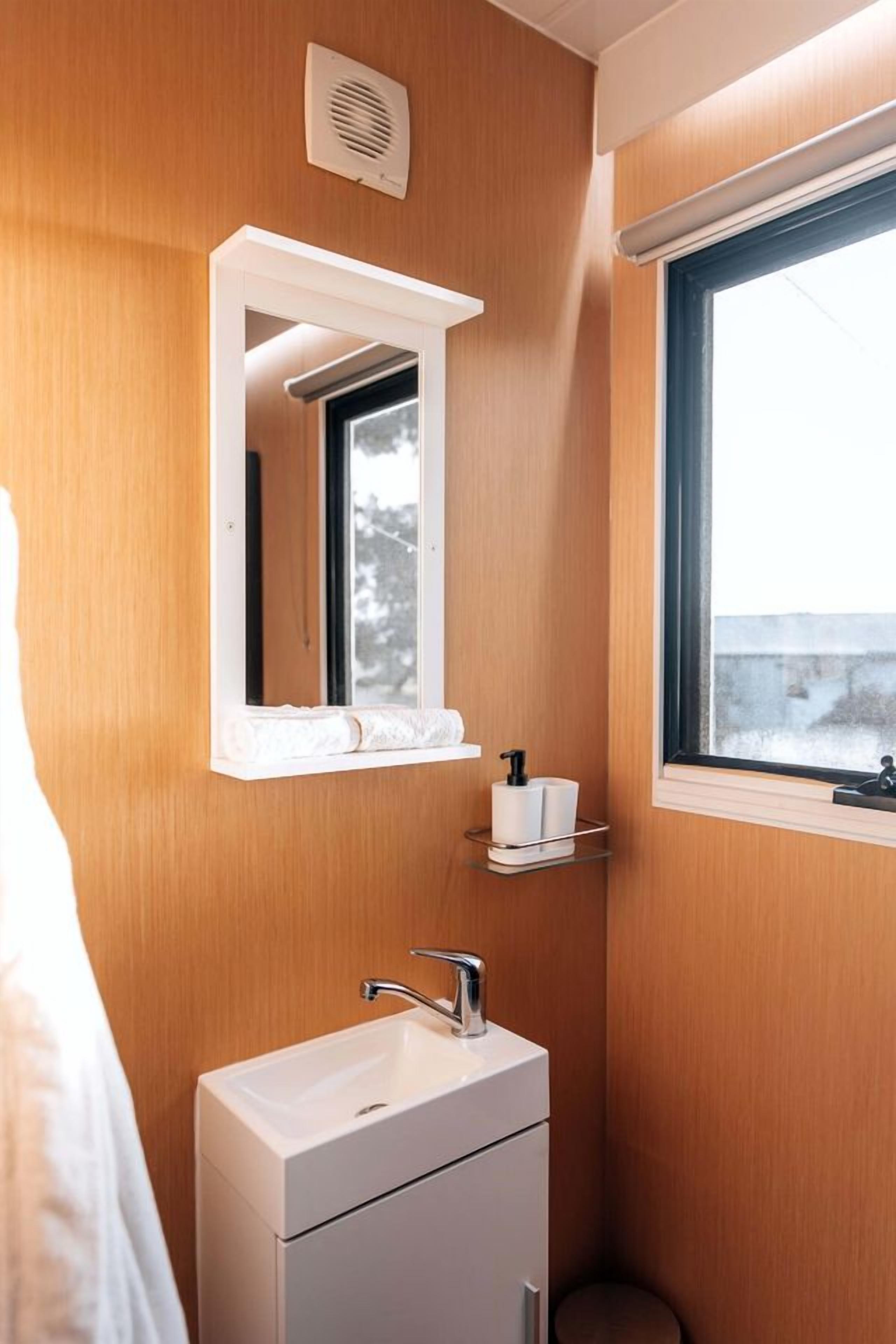 A bathroom with a wooden wall, a white sink with a faucet, a mirror, a window, and a toilet paper holder.