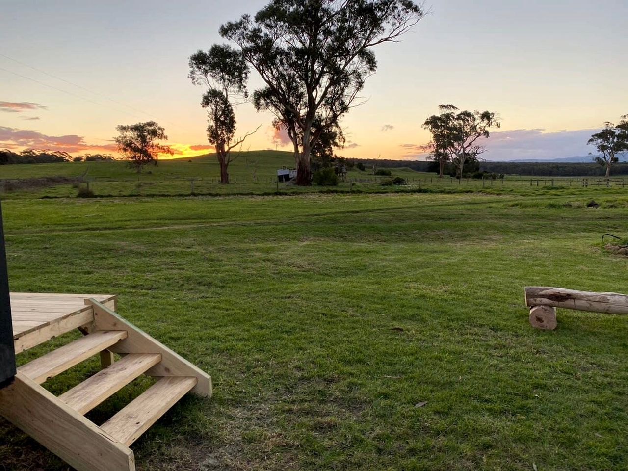 A grassy field with a wooden staircase leading up to a building, surrounded by trees and a sunset in the background.
