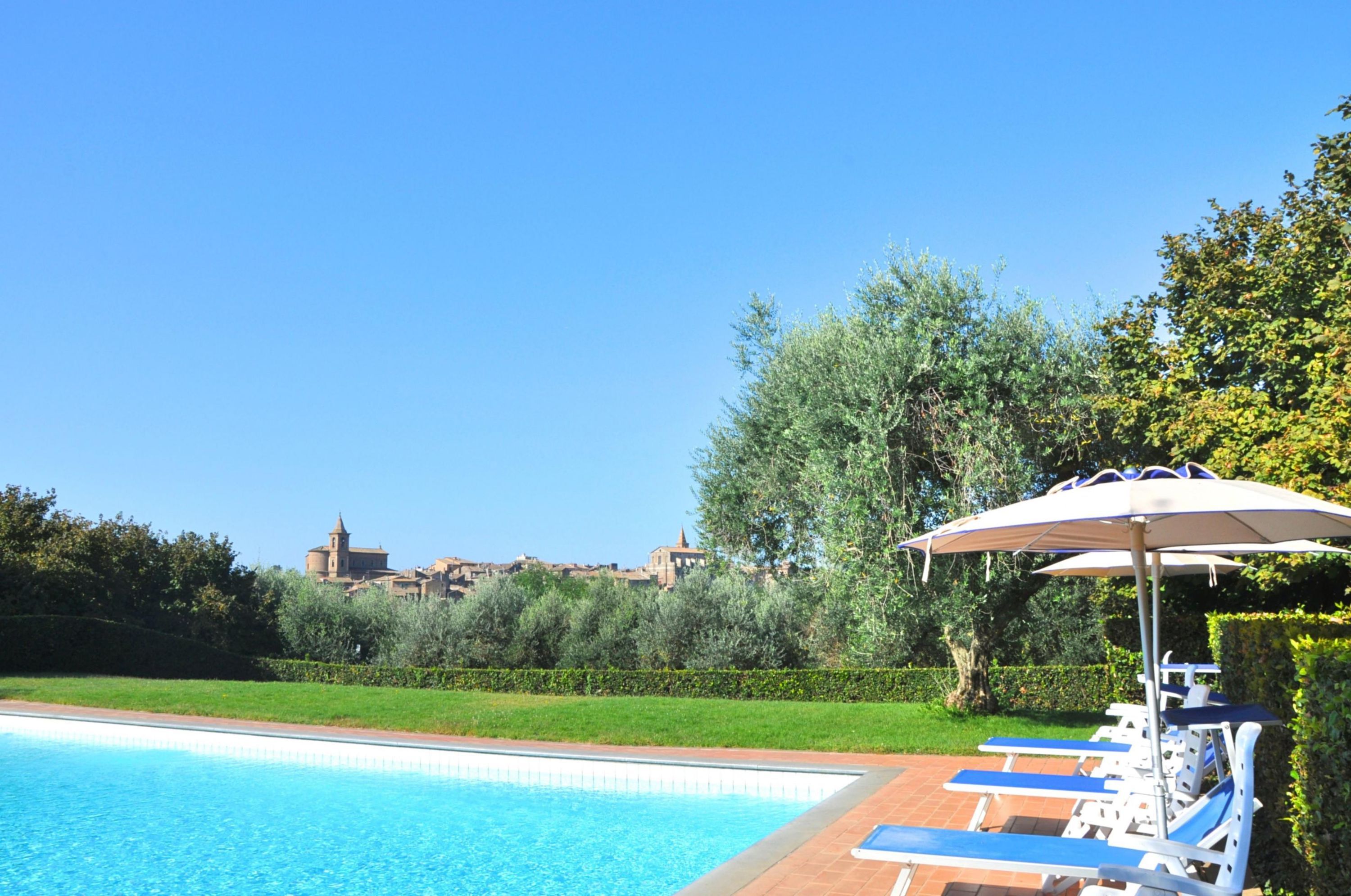 A swimming pool with lounge chairs and an umbrella, surrounded by greenery and a view of a historic building.