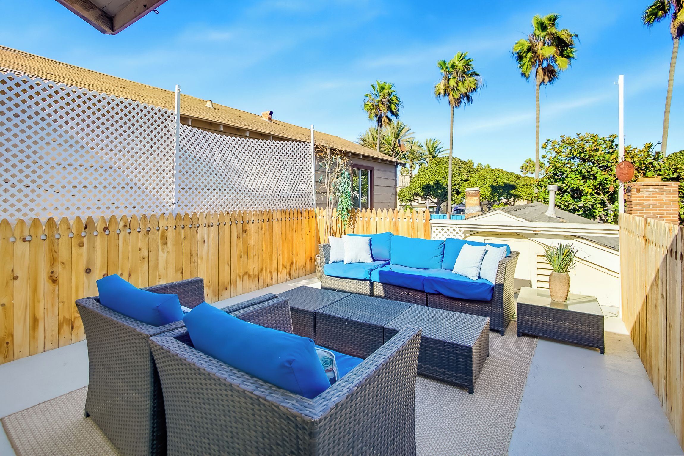 Un espace extérieur aménagé en terrasse, avec des meubles en osier, une clôture en bois et des palmiers en arrière-plan.
