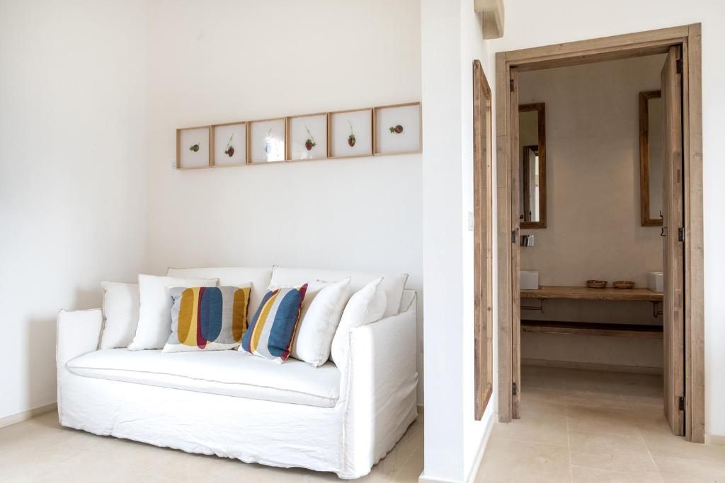 A white sofa with colorful cushions, a wall-mounted picture frame with fruit, and a wooden door leading to a bathroom.