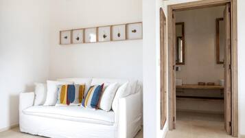 A white sofa with colorful cushions, a wall-mounted picture frame with fruit, and a wooden door leading to a bathroom.