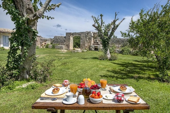 Une table de pique-nique installée à l’extérieur, avec de la nourriture et des boissons, entourée de verdure et avec des vestiges historiques en arrière-plan.