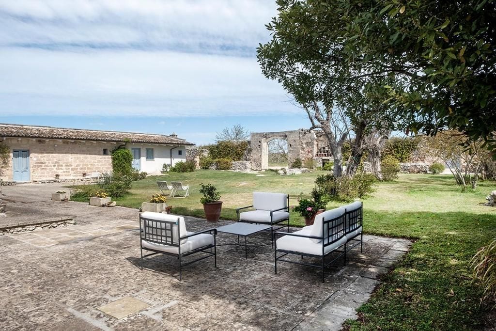 Un patio con mobiliario exterior, un edificio de piedra y una zona verde con árboles.