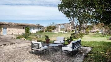 Un patio con mobiliario exterior, un edificio de piedra y una zona verde con árboles.