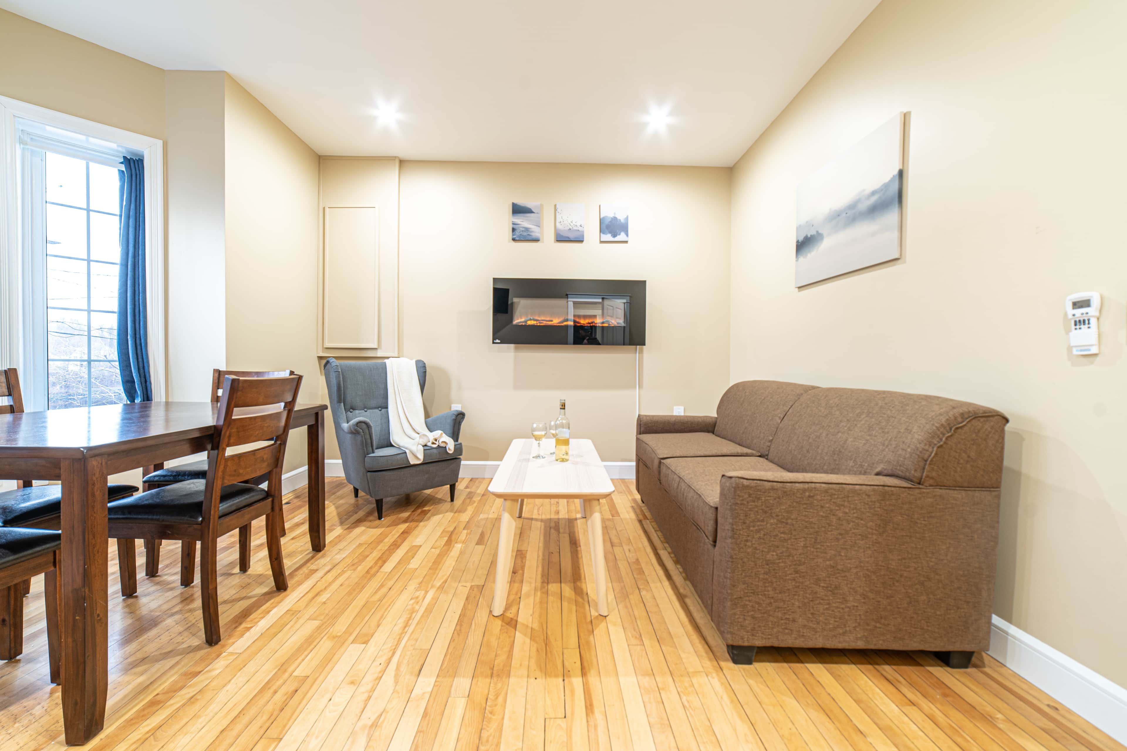 A living room with a wooden floor, a brown sofa, a dining table with chairs, a fireplace, and a TV mounted on the wall.