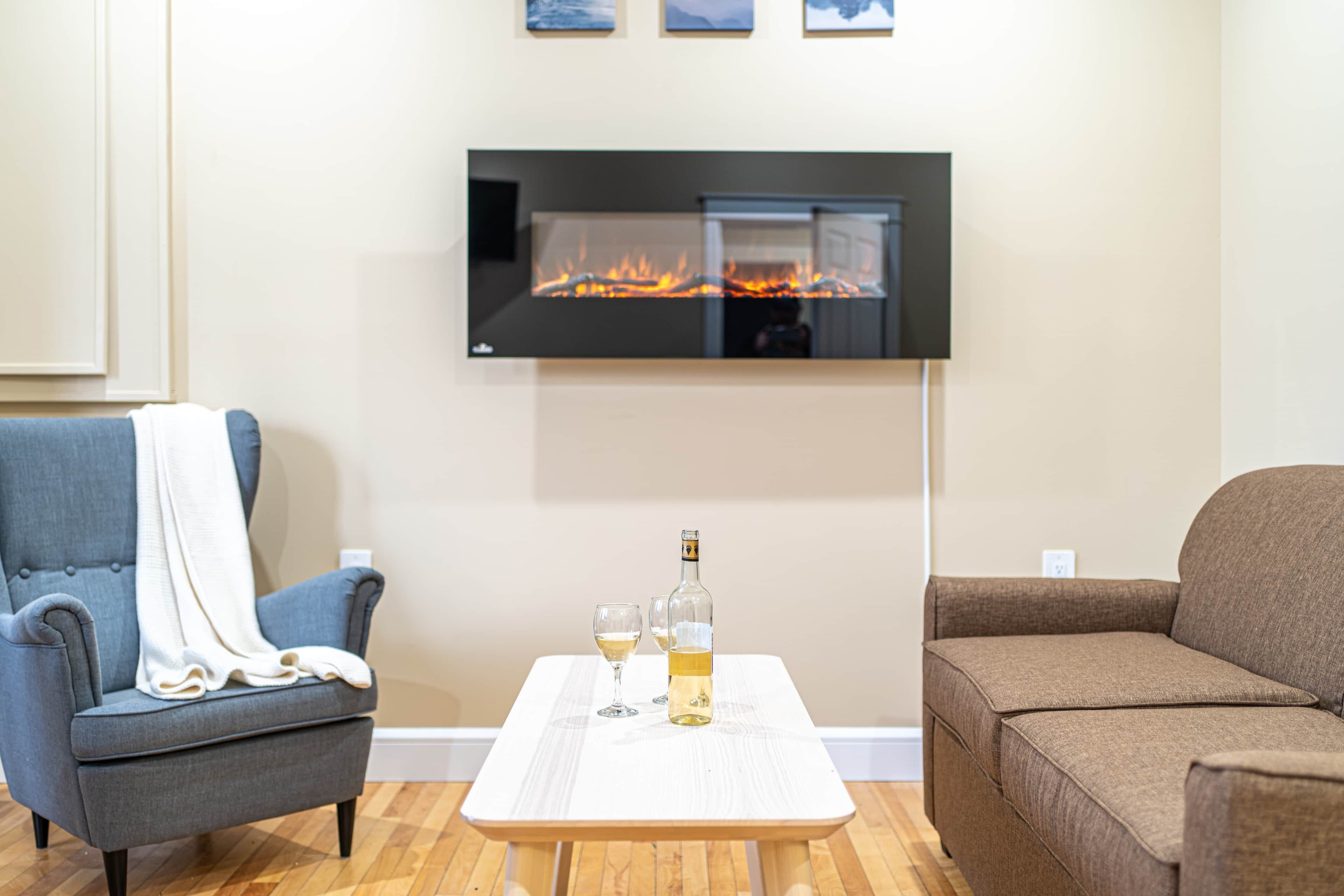 A living room with a flat-screen TV mounted on the wall, a fireplace, a blue armchair, a brown sofa, and a small table with glasses and a bottle.